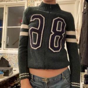 Grön stickad tröja med nummer 28 - Säljer en supercool grön stickad tröja.  Tröjan har långa ärmar med vita ränder och en dragkedja framtill. Perfekt för höst och vinter, och den är både varm och stilren. ❤️