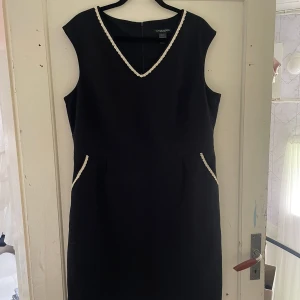 Svart klänning från Ellos - Säljer en elegant svart klänning från Ellos, perfekt för festliga tillfällen. Klänningen har en v-ringad hals och är ärmlös, vilket ger en stilren look. Den har fina detaljer med vita kanter vid halsen och fickorna. Passar perfekt för både höst och vinter.