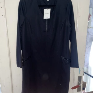 Svart klänning från Cellbes - Säljer en elegant svart klänning från Cellbes i storlek 46/48. Klänningen har en v-ringad design och långa ärmar, perfekt för både vardag och fest. Den har dragkedjedetaljer vid fickorna som ger en modern touch.