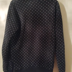 Svart tröja med prickigt mönster från SuperDry - Säljer en svart tröja från SuperDry med ett coolt vitt prickigt mönster över hela. Den har långa ärmar och en stor logga på framsidan. Perfekt för en avslappnad stil!