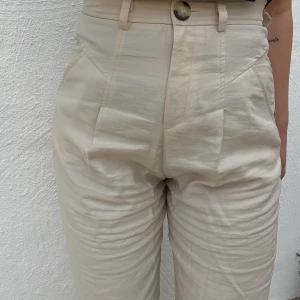 Beige linnebyxor - Säljer ett par snygga beige linnebyxor. De är högmidjade med en knapp och dragkedja framtill. Byxorna har en avslappnad passform och är perfekta för både vardag och sommarhäng. De har också praktiska fickor på sidorna. Aldrig använda
