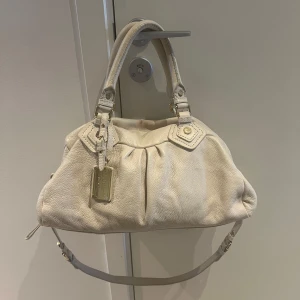 Marc Jacobs väska  - Säljer en snygg beige handväska i skinn med dubbla handtag och en justerbar axelrem. Väskan har en dragkedja upptill och ett coolt mönstrat innerfoder. Perfekt för både vardag och fest! Det finns en liten fläck på ena sidan, men annars i bra skick. Skriv om ni har fler frågor🩷 Kan även möta upp i centrala Stockholm📍
