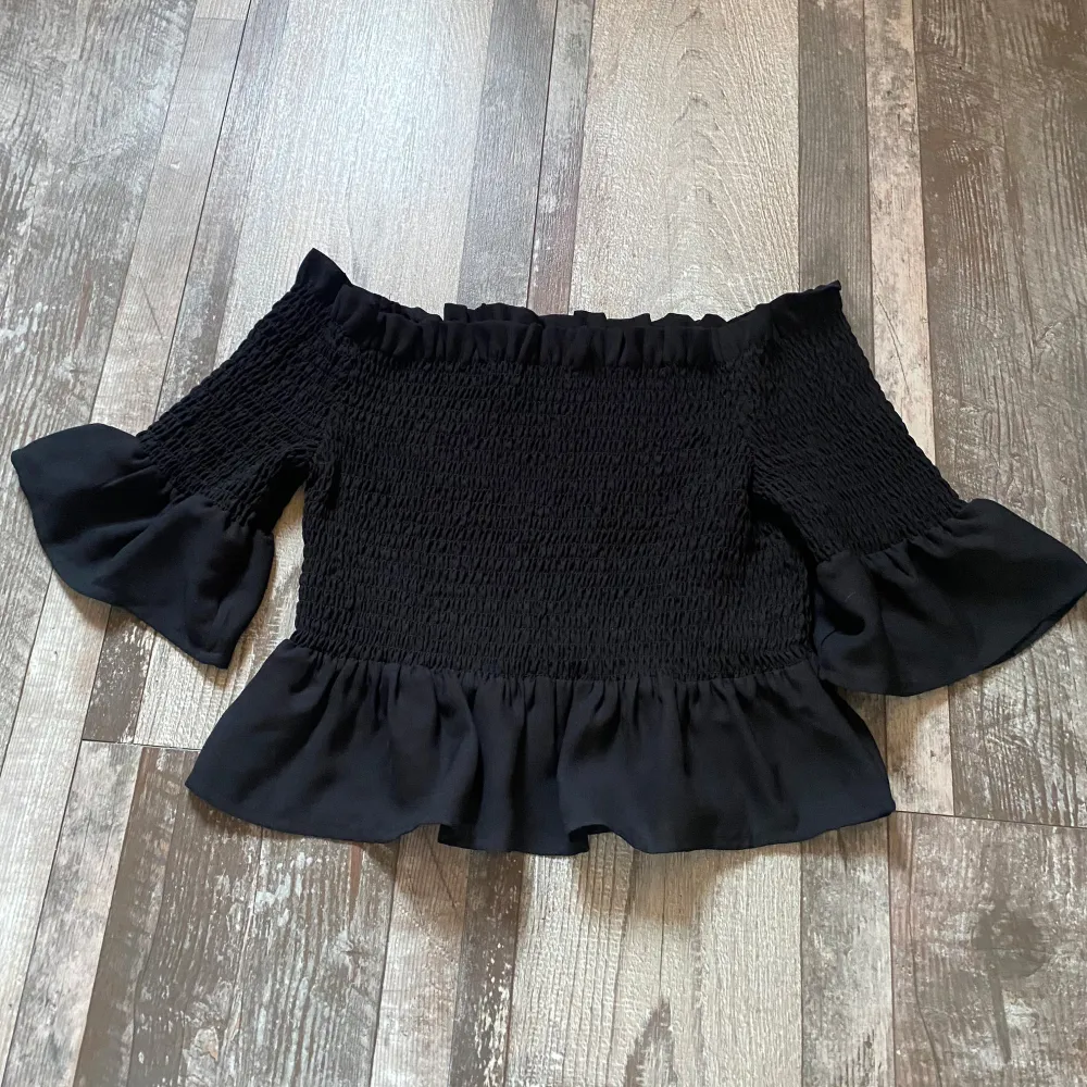 Säljer en superfin svart offshoulder topp med volanger. Den har en smockad design som ger en tight passform och vida ärmar med volangdetaljer. Perfekt för festliga tillfällen eller en utekväll. Toppen är i mycket bra skick och redo för ett nytt hem! (plagget tvättas innan det fraktas). Toppar.