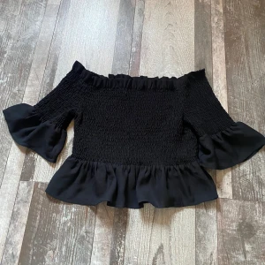 Svart offshoulder topp med volanger - Säljer en superfin svart offshoulder topp med volanger. Storlek L men sitter som S-L. Den har en smockad design som ger en tight passform och vida ärmar med volangdetaljer. Perfekt för festliga tillfällen eller en utekväll. Toppen är i mycket bra skick och redo för ett nytt hem! 