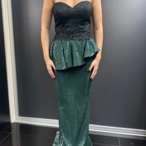 Svart och grön axelbandslös klänning - Snygg axelbandslös klänning i svart och grön med glittrig finish. Den har en volangdetalj i midjan och en lång, elegant kjol. Perfekt för festliga tillfällen!