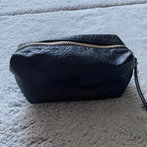 Svart necessär med dragkedja - Säljer en stilren svart necessär i skinn med en praktisk dragkedja. Perfekt för att hålla ordning på dina småsaker när du är på språng. Den har en enkel design och är lätt att bära med sig. Perfekt för resor eller som en snygg accessoar i badrummet. Ca 19 cm bred.