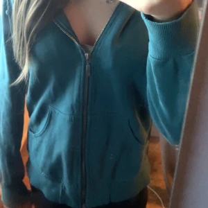 Grön huvtröja från Lindex - Säljer en snygg grön huvtröja från Lindex. Den har dragkedja framtill och en skön huva med snören. Perfekt för en avslappnad stil och passar bra till både höst och vår. Tröjan har långa ärmar och är i bra skick. 