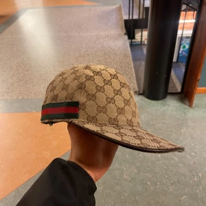 Beige keps från Gucci - Säljer en snygg beige keps från Gucci med deras ikoniska monogrammönster. Kepsen har en grön och röd randdetalj på sidan och justerbar rem baktill. Perfekt för att ge din outfit en lyxig touch.!     Köptes i 2023 vår kvitto finna måste ba leta fram pris kan diskuteras!!