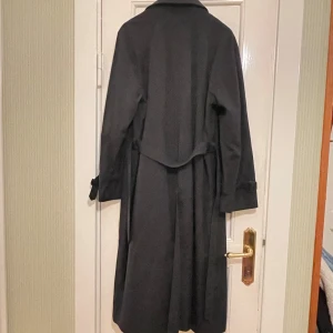 Svart lång kappa - Säljer en elegant svart lång poplinkappa i mycket bra skick. Perfekt för höst och vår med sin klassiska stil och långa ärmar. Kappan har knappar framtill, skärp i midjan och en stilren krage. Passar både till vardags och festliga tillfällen. Ett måste i garderoben för den stilmedvetna! Längd 112 cm.