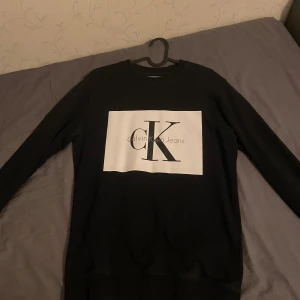 Svart sweatshirt från Calvin Klein - Säljer en stilren svart sweatshirt från Calvin Klein med deras ikoniska logga i vitt på bröstet. Tröjan har långa ärmar och är perfekt för en casual look. Passar både till vardags och lite finare tillfällen. Superbekväm och lätt att matcha med andra plagg!