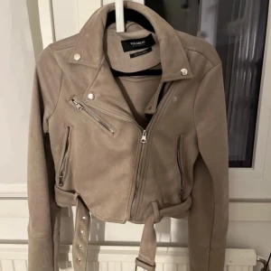 Beige mockajacka från Pull&Bear - Säljer en snygg beige mockajacka från Pull&Bear. Jackan har en cool bikerstil med dragkedjor och ett bälte i midjan. Perfekt för höst och vår! Den är i mycket bra skick och har långa ärmar. Passar perfekt för en casual look eller en utekväll.