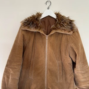 Brun manchesterjacka med fuskpäls - Säljer en snygg brun manchesterjacka med fuskpälsdetaljer på huva och ärmslut. Jackan har en dragkedja framtill och är perfekt för höst och vinter. Den är i bra skick och supermysig att ha på sig när det är kallt ute. Perfekt för att hålla stilen i kylan!