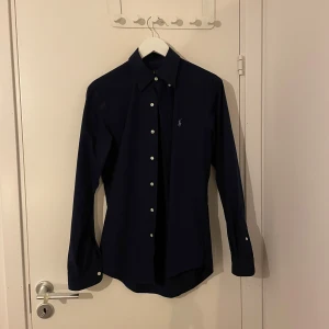 Mörkblå skjorta från Ralph Lauren - Säljer en stilren mörkblå skjorta från Ralph Lauren i custom fit. Skjortan har långa ärmar och klassiska knappar framtill. Perfekt för både vardag och festliga tillfällen. Passar bra till både jeans och kostymbyxor. Skjortan är i mycket bra skick och redo för nya äventyr!