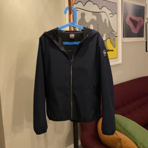 Marinblå vindjacka med huva - Säljer en snygg marinblå vindjacka från Colmar. Jackan har en praktisk huva och dragkedja framtill. Perfekt för höst och vår, med en stilren design som passar till det mesta. Den är i mycket bra skick och redo för nya äventyr!