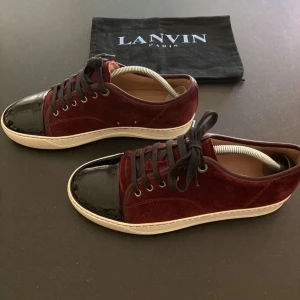 Vinröda sneakers från Lanvin - Snygga vinröda sneakers från Lanvin med svarta lackdetaljer på tån och snörning. Skorna har en stilren design med en vit sula som ger en modern touch. Skicket är mycket bra med inga defekter. För fler bilder eller frågor kom privat. Priset ej hugget i sten!