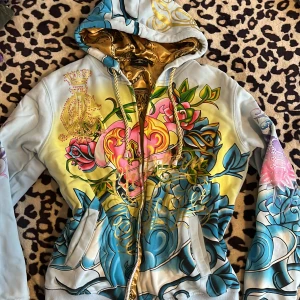 Färgglad hoodie från Christian Audigier - Säljer en unik huvtröja från Christian Audigier med färgglada blommönster och detaljer i guld. Tröjan har en dragkedja framtill och en fodrad huva med mönster. Perfekt för dig som vill sticka ut med en cool och konstnärlig stil.