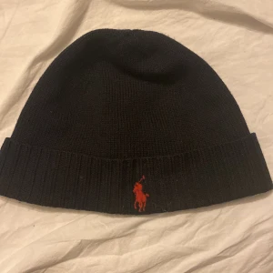 Mörkblå mössa från Polo Ralph Lauren - Snygg mörkblå mössa från Polo Ralph Lauren i 100% ull. Den har en ribbad kant och är dekorerad med ett rött broderat logotypmotiv. Perfekt för att hålla värmen med stil.