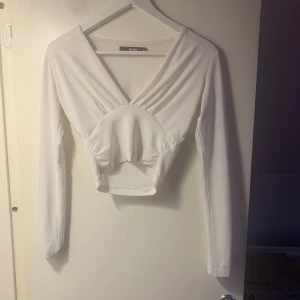 Vit långärmad topp från Bik Bok - Säljer en stilren vit långärmad topp från Bik Bok i storlek XS. Toppen har en v-ringad design med en unik detalj framtill som ger en snygg look. Perfekt för att matcha med både jeans och kjol.