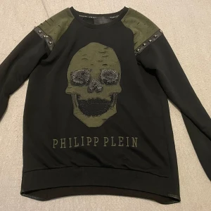 Svart härlig tröja  från Philipp Plein - Snygg svart tröja från Philipp Plein med en stor dödskalle på framsidan. Tröjan har långa ärmar och coola detaljer med nitar på axlarna. Perfekt för vintern.