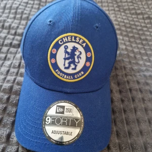 Blå Chelsea keps från New Era - Snygg blå keps från New Era med Chelsea Football Club-logga framtill. Kepsen är justerbar och har en klassisk design med böjd skärm. Perfekt för fotbollsfans!