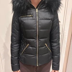 Svart dunjacka från Rockandblue i storlek 32 - Snygg svart dunjacka från Rockandblue med pälskantad huva och guldfärgade dragkedjor. Jackan har en quiltad design och flera praktiska fickor. Perfekt för kyliga dagar. Jackan köptes för  2 200 kronor. Knappt använda då storleken blev snabbt liten :)