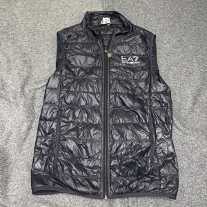 Svart väst från Emporio Armani - Snygg svart quiltad väst från Emporio Armani med dragkedja och logotyp på bröstet. Perfekt för lager-på-lager under kyliga dagar.