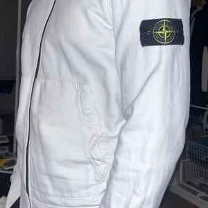 Vit jacka från Stone Island - Snygg vit jacka från Stone Island med dragkedja och huva. Hoodie har en ikonisk patch på ärmen och praktiska fickor framtill. Perfekt för en stilren look.