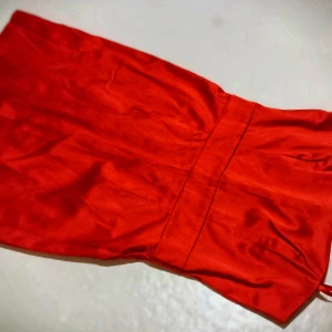 Röd axelbandslös klänning - Snygg röd axelbandslös klänning i satin med dragkedja baktill. Perfekt för festliga tillfällen med sin eleganta och stilrena design.