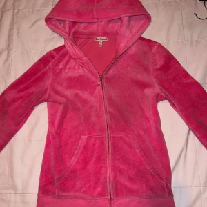 Rosa hoodie från Juicy Couture - Säljer en snygg rosa hoodie från Juicy Couture med dragkedja och huva. Den har ett stort, glittrigt JC-märke på ryggen med paljetter. Perfekt för en avslappnad och trendig look.