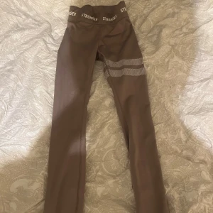 Bruna leggings från Stronger - Snygga bruna leggings från Stronger med elastisk midja och textdetaljer. Perfekta för träning eller en avslappnad dag. Tillverkade i ett stretchigt material för optimal komfort.