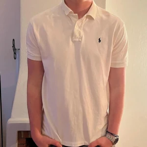 Vit pikétröja från Ralph Lauren - Säljer en klassisk vit pikétröja från Ralph Lauren med korta ärmar och den ikoniska loggan på bröstet. Perfekt för en stilren och avslappnad look. Perfekt nu att klippa till så här innan nyår. Skriv för frågor, piken är sparsamt använd men ni ser själva skicket på den