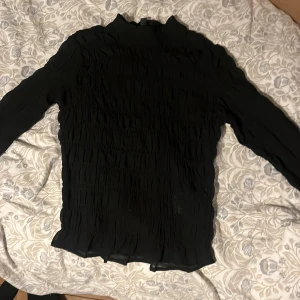 Svart långärmad tröja från H&M Divided - Säljer en svart långärmad tröja från H&M Divided. Tröjan har en unik rynkad textur som ger den ett stilrent utseende. Perfekt för att lägga till en touch av elegans till din garderob.