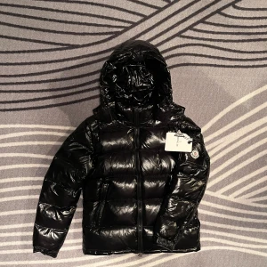 Svart pufferjacka från Moncler - Snygg svart pufferjacka från Moncler med glansig finish och huva. Jackan har en dragkedja framtill och Moncler-logga på ärmen. Perfekt för kyliga dagar.