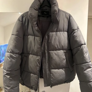 Grå pufferjacka - Säljer en stilren grå pufferjacka med dragkedja. Jackan har en hög krage och är perfekt för kyligare dagar. Den quiltade designen ger extra värme och komfort. Normal i storleken. Använd få gånger som nyskick. Nypris 600