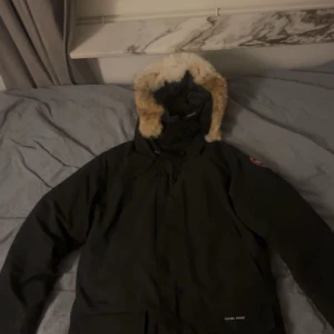 Canada Goose Langford Parka Heritage - Säljer en svart dunjacka från Canada Goose med pälskantad luva. Jackan är quiltad och har långa ärmar, perfekt för kalla vinterdagar. Den har en klassisk design med dragkedja och flera praktiska fickor.                                                                            Väldigt bra skick, inga skador. Kvitto finns! Hör av dig om du har frågor!