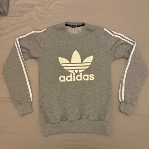 Grå sweatshirt från Adidas - Säljer en grå sweatshirt från Adidas med det klassiska trefoil-logotypen på framsidan. Tröjan har långa ärmar med de ikoniska tre ränderna längs ärmarna. 