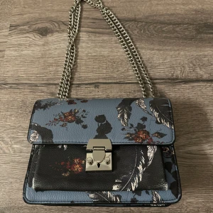 Blommig axelväska med kedja - Snygg axelväska i blått med blommigt mönster och fjäderdetaljer. Väskan har en stilren kedja som axelrem och ett metallspänne framtill. Perfekt för att ge din outfit en unik touch.