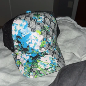 Blommig keps  - Trendig keps med blommigt mönster i blått och grönt. Har en justerbar rem med röd och grön rand och svart nätpanel baktill för ventilation.