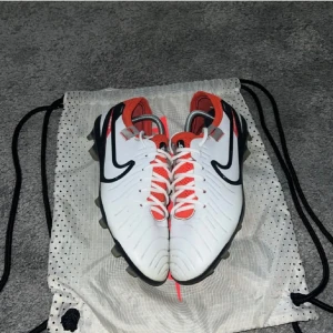 Nike Tiempo fotbollsskor - Säljer ett par Nike Tiempo fotbollsskor i vitt med svarta och röda detaljer. Skorna har snörning och en robust sula för bra grepp på planen. Perfekta för fotbollsspelare som söker stil och funktion.
