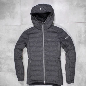 Grå dunjacka från Peak Performance - Snygg grå dunjacka från Peak Performance med huva och dragkedja. Perfekt för kyliga dagar. Jackan har en stilren design med långa ärmar och diskreta logodetaljer på bröstet och ärmen. PRIS KAN DISKUTERAS! 