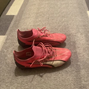 PUMA ULTRA ULTIMATE FG/AG PHENOMENAL - POISON PINK/VIT/SVART - Säljer ett par rosa fotbollsskor från Puma med snörning och svarta detaljer. Skorna har en strömlinjeformad design och är perfekta för fotbollsplanen. De har en robust sula med dobbar för optimalt grepp.
