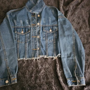 Croppad jeansjacka med slitna detaljer - Snygg croppad jeansjacka i blått med slitna detaljer och knappar framtill. Jackan har två bröstfickor med knapp och fransig nederkant för en trendig look. Perfekt för en avslappnad stil.