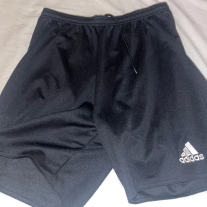 Svarta träningsshorts från Adidas - Säljer ett par svarta träningsshorts från Adidas. De har en elastisk midja med dragsko för justerbar passform och Adidas-loggan på benet. Perfekta för sport och träning.