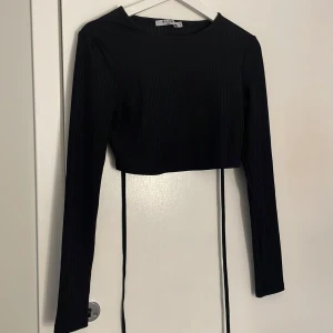 Svart ribbad croppad topp - Snygg svart ribbad och croppad topp med långa ärmar och knytning i ryggen. Från NAKD. Stretchig 