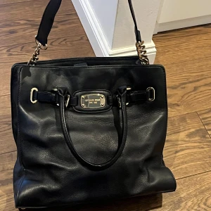 Svart skinnväska från Michael Kors - Snygg svart skinnväska från Michael Kors med gulddetaljer och dubbla handtag. Väskan har en elegant design med en metallplatta framtill och kedjedetaljer på sidorna. Perfekt för att bära dina dagliga nödvändigheter med stil.                                   Skriv om du vill ha fler bilder! Köparen tar hand om frakt😊