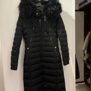 Svart lång dunjacka med pälskrage - Snygg svart lång dunjacka från cedrico med en fluffig pälskrage. Jackan har dragkedja och knappar framtill samt praktiska fickor med knappar. Perfekt för kalla vinterdagar. Svart tvättbjörnspäls