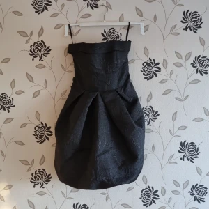 Svart glittrig klänning från Atmosphere, Strl: 36/S, nyår - Elegant svart klänning från Atmosphere med axelbandslös design och en stor rosett baktill. Klänningen har en unik struktur och voluminös kjol som ger en stilfull look. Perfekt för speciella tillfällen.