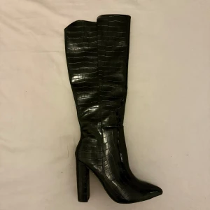 Svarta stövlar med krokodilmönster - Snygga svarta stövlar med krokodilmönster och hög klack. De har en spetsig tå och dragkedja på sidan för enkel på- och avtagning. Perfekta för att ge en stilren touch till din outfit.
