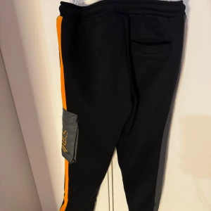 Svarta mjukisbyxor från Jack & Jones 164 - Snygga svarta mjukisbyxor från Jack & Jones med orangea detaljer och en cool ficka på sidan. Byxorna har snörning i midjan för bästa passform och är perfekta för en avslappnad stil.