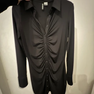 Svart klänning från H&M - Snygg svart klänning från H&M med långa ärmar och knappar framtill. Klänningen har en stilren design med rynkade detaljer längs knäppningen som ger en unik touch. ANVÄND EN GÅNG!! Sitter tight på kroppen!